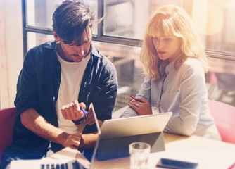 Wall Mural - Two young coworkers working on laptop computer at sunny office.Woman holding paper documents and pointing on notebook screen. Horizontal.Blurred background.Cropped