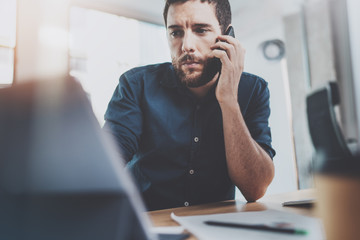 Bearded young businessman working at sunny office.Man using contemporary smartphone.Horizontal.Blurred background.Flares.