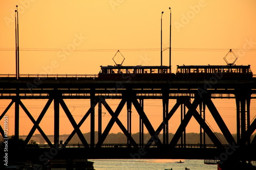 Fotomural Siluetas de un puente de dos niveles y 2 tranvías en una puesta de sol, Dnepr ci