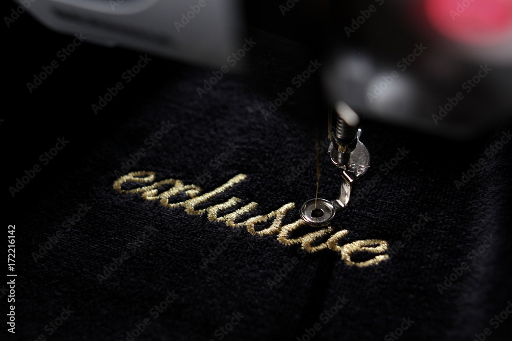 embroidery of gold lettering "exclusive" on black velvetely fabric with embroidery machine - diagonal view with part of machine - background and foreground blanked out blurry  - obrazy, fototapety, plakaty 