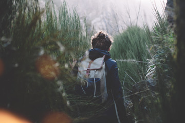 Young girl walking among huge grass in mountains with backpack in jacket. Cool weather.