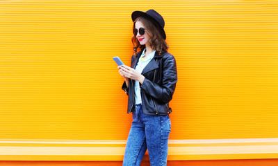 Fashion smiling woman is using smartphone in black rock jacket on colorful orange background