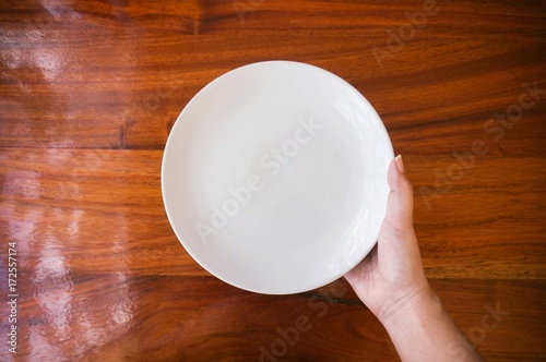 Fotografie, Obraz Female (woman) hands hold (support) a white dish (plate) on wooden table