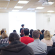 © kasto - Business and entrepreneurship symposium. Female speaker giving a talk at business meeting. Audience in conference hall. Rear view of unrecognized participant in audience. Copy space on whitescreen.