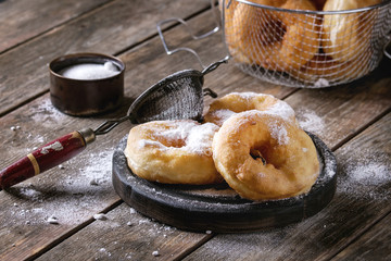 Homemade donuts with sugar powder on black serving board and in frying basket served with vintage sieve on old wooden plank table. Rustic style.