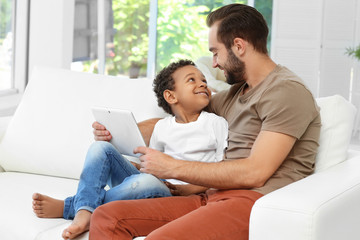 Happy father with adopted African-American boy using tablet computer while sitting on couch at home