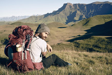 Male Hiker With His Back Pack Taking A Break In A Field Surrounded By Mountains.