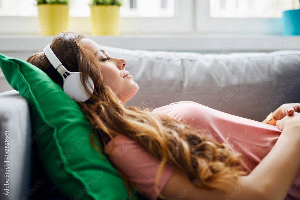 Portrait of a beautiful young woman lying on sofa with headphones on and closed eyes, relaxing - obrazy, fototapety, plakaty 