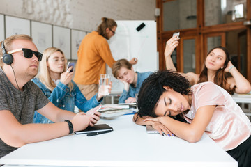 Wall Mural - Young teacher writing on board while students sitting at the table and aren't listening him during lesson. Group of students making photos,listening music and sleeping while teacher isn't seeing it