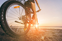 A Woman In A Multicolored Suit Sits On A Bicycle In A Desert Area Near The Water. Fitness Concept. Rear View And Bottom View. Close-up