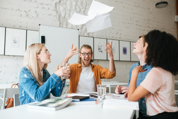 Wall Mural - Portrait of young emotional boy with blond hair and beard happily throw up papers while sitting with friends. Group of people working together. Students studying with books in classroom