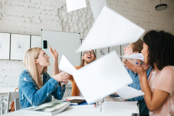 Wall Mural - Portrait of emotional people happily throw up papers in office while working together. Group of young students joyfully studying with books on table in classroom.