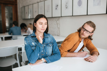 Wall Mural - Portrait of two students sitting at the desk in classroom and writing test. Young man in glasses thoughtfully looking at girl near and closing by hand his test while she carefully trying write off