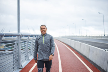 Cheerful young dark-skinned sportsman in stylish outfit walking along rubber covered surface of road, feeling excited about running marathon. People, sports, determination and active lifestyle