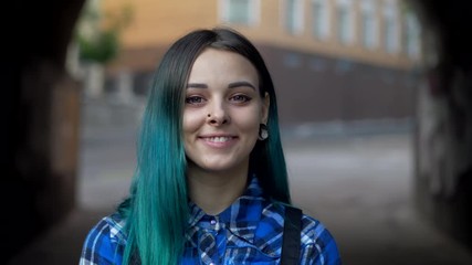 Wall Mural - Street trendy or hipster girl with blue dyed hair. Woman with piercing in nose, violet lenses, ears tunnels and unusual hairstyle stands in city. 4k