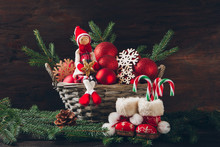 Doll Sitting In A Basket With Fir Tree Branches, Red Balls And Snowflakes.