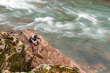 hobby, high qualification, nature concept. professional photographer is at work, standing with tripod on the rocky coast nearby with river with choppy waters that flowing away very fast