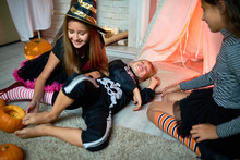 Playful Cute Little Witch Tickling Foot Of Boy Dressed As Skeleton Lying On Floor, Smiling Friend Observing Them At Halloween Party