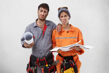 Young overworked male and female builders look with gloomy expressions, have no desire to work any more, hold sketches and drawings, want to rest for minute. Unwillingness and tiredness concept