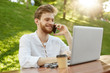 © Cookie Studio - Young beautiful redhead male freelancer with good haircut and beard, working on laptop in park,talking on phone with boss and feeling happy about job achievements