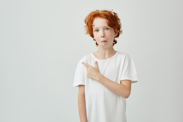 Wall Mural - Funny ginger boy with freckles having curious expression and pointing a side with finger on white wall. Copy space for advertisement.