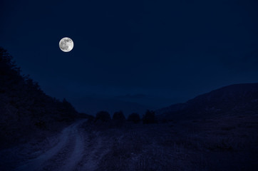 Mountain Road through the forest on a full moon night. Scenic night landscape of dark blue sky with moon