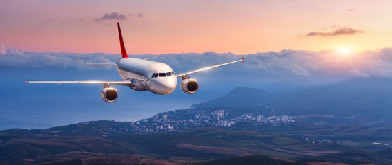 Passenger airplane. Landscape with white airplane is flying in the orange sky with clouds over mountains, sea at colorful sunset. Passenger aircraft is landing. Commercial plane. Private jet. Travel