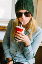 Stylish Young Woman Wearing Sunglasses Sitting In Restaurant Drinking Soda Through Straw
