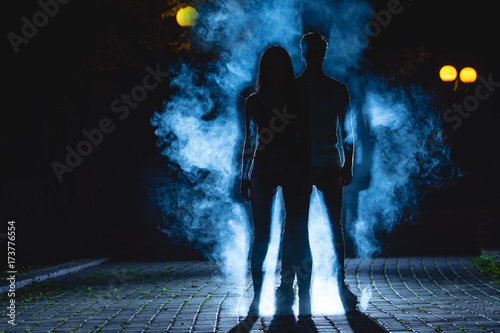Fotografía The man and woman stand on a dark alley on the blue fume background
