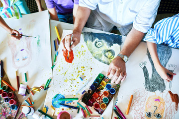 Top view of childrens drawings on table in art class with group of people painting and teachers hands