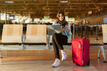 Airport Young female passenger on smart phone and laptop sitting in terminal hall while waiting for her flight