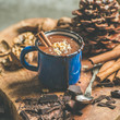Rich winter hot chocolate with cinnamon sticks and walnuts in blue enamel mug on wooden board over grey concrete background, selective focus, square crop
