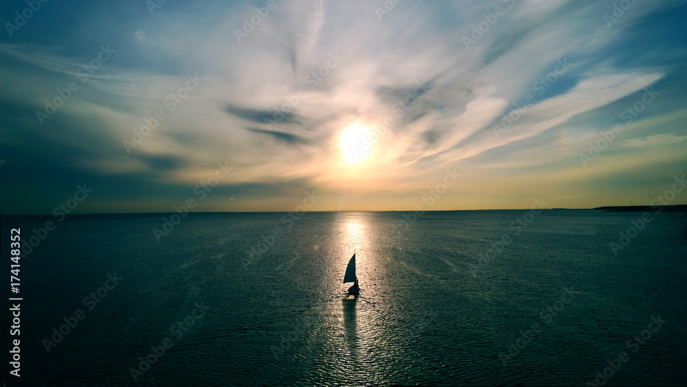 Little white boat floating on the water towards the horizon in the rays of the setting sun. Beautiful clouds with yellow highlights. Aerial view - obrazy, fototapety, plakaty 