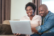 © mavoimages - Laughing African couple sitting on a sofa using a laptop