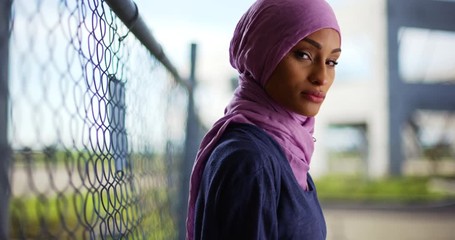 Wall Mural - Portrait of black female in purple headscarf looking at camera with serious expression. Muslim woman standing by chain-link fence, looking at camera with confidence 