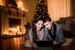 © MestoSveta - A cozy evening of a young family watching something together on a tablet, christmas decorations around in their room. Caucasian Couple using digital tablet at home celebrating New Year.
