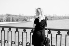 Attractive American Blonde Hair Woman Walk In The City Wear In Small Black Dress And Bag. Lady On A Bridge In European Town