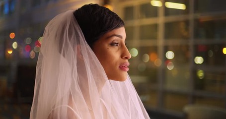 Canvas Print - Close up of beautiful bride in white wedding veil standing on city street in evening smiling. Profile of young black female with gentle smile standing in urban setting at night 
