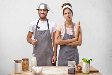 Wall Mural - Tired attractive female cook work all day, holds rolling pin, stands next to her male colleague who is full of energy, going to bake pie or whisking cream, has good mood and pleased expression.