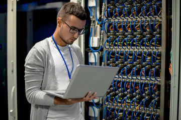 Side view  portrait of young man  using  laptop standing by server cabinet while working with supercomputer