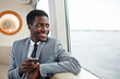 Happy African-american businessman with smartphone sitting by steamer window and looking through it