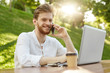 © Cookie Studio - Close up of cheerful red-haired guy with trendy hair and beard sitting outside, working on laptop computer, talking on phone with best friend about startup project.