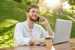 © Cookie Studio - Portrait of young successful european ginger man with stylish hairstyle and beard in white shirt sitting in park, smiling. working on his laptop, drinking coffee and talking on phone with his partner.