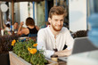 © Cookie Studio - Beautiful ginger male freelancer with stylish haircut and beard smiles, looking at camera and working on laptop computer outside cafeteria.