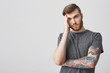 Close up of beautiful unhappy bearded hipster man with tattoo and good hairstyle holding head with hand, looking in camera with tired expression being exhausted after first day at work.
