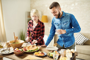 Portrait of modern young couple  standing at festive dinner table  cutting food and smiling at gathering with friends