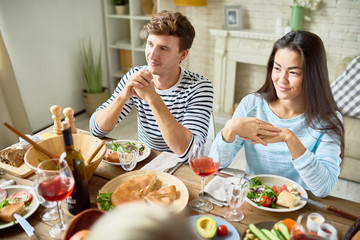 Portrait of two modern young people sitting at big table with food having dinner with friends and smiling happily celebrating holiday