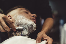 Crop Barber Using Razor And Shaving Tattooed Man In Chair.