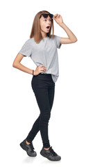 Wall Mural - Surprised teen girl in full length looking with opened mouth away at blank copy space raising her sunglasses in astonishment, isolated on white background