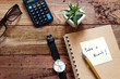Wood Desktop work space have brown glasses,calculator,pot plant ,black watch,pen,notebook,and note pad with 